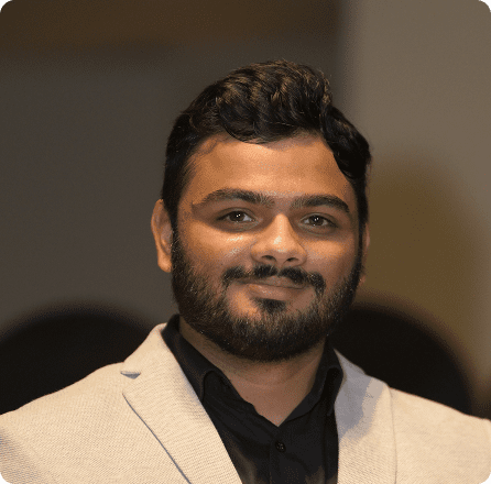 A man with a beard smiles confidently, wearing a light gray suit jacket over a black shirt.