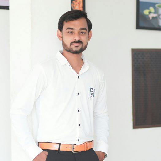 A young man in a white shirt and black trousers leans against a wall, smiling confidently in a modern interior space.